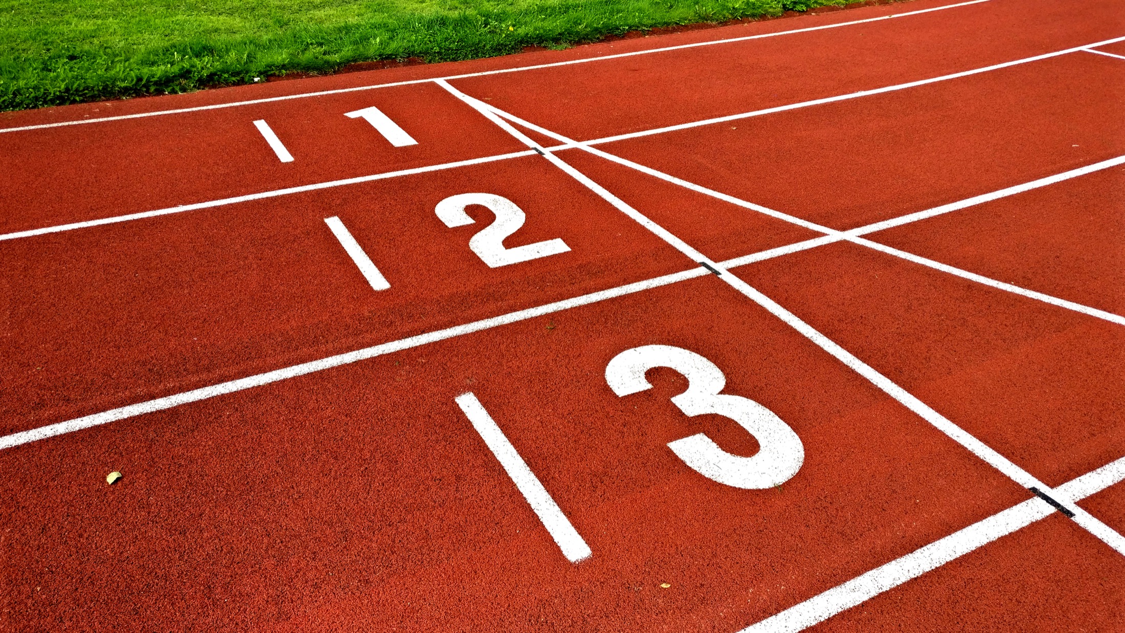 A running track with lanes one, two, and three marked in white on an orange all-weather surface, bordered by green grass.